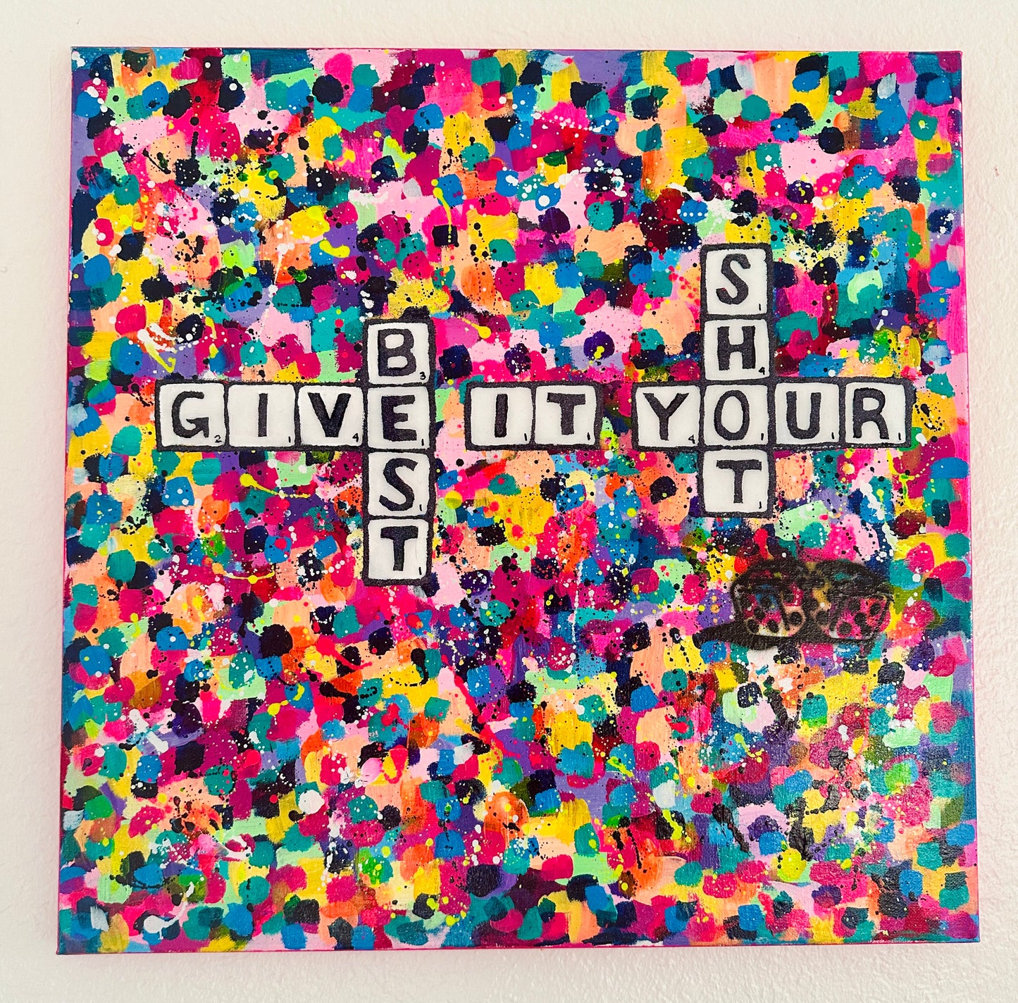 Colorful abstract splatter painting with black-and-white Scrabble-style tiles reading “Give It Your Best Shot” on canvas